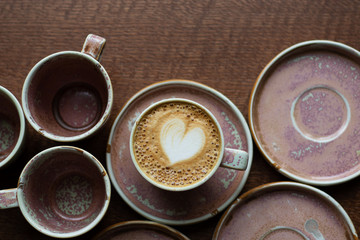 A cup of coffee in the shape of a heart with milk foam. Cappuccino among the dishes on the table. A service of one color from mugs and saucers. Top View