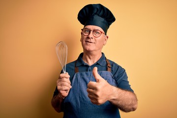 Middle age hoary cooker man wearing apron and hat holding whisk over yellow background happy with big smile doing ok sign, thumb up with fingers, excellent sign