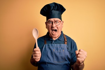 Naklejka premium Middle age cooker man wearing apron and hat holding wooden spoon over yellow background screaming proud and celebrating victory and success very excited, cheering emotion