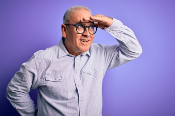 Middle age handsome hoary man wearing striped shirt and glasses over purple background very happy and smiling looking far away with hand over head. Searching concept.
