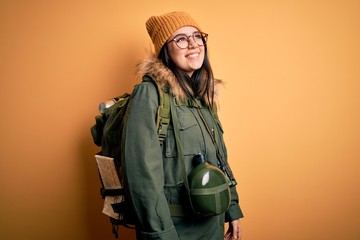 Young hiker woman wearing hiking backpack, canteen and map over yellow background looking away to side with smile on face, natural expression. Laughing confident.