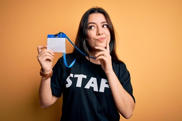 Young brunette worker woman wearing staff t-shirt as uniform showing id card serious face thinking...