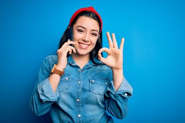 Young brunette woman talking on smartphone over blue isolated background doing ok sign with fingers, excellent symbol