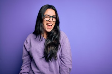 Young brunette woman wearing glasses over purple isolated background winking looking at the camera...