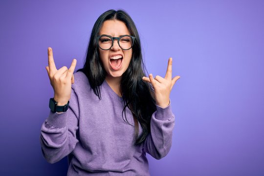 Young brunette woman wearing glasses over purple isolated background shouting with crazy expression doing rock symbol with hands up. Music star. Heavy concept.