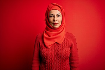 Middle age woman wearing traditional muslim hijab standing over isolated red background with serious expression on face. Simple and natural looking at the camera.