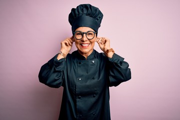 Middle age brunette chef woman wearing cooker uniform and hat over isolated pink background Smiling pulling ears with fingers, funny gesture. Audition problem