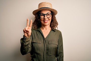 Middle age brunette woman wearing glasses and hat standing over isolated white background showing and pointing up with fingers number two while smiling confident and happy.