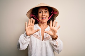 Middle age brunette woman wearing asian traditional conical hat over white background afraid and terrified with fear expression stop gesture with hands, shouting in shock. Panic concept.