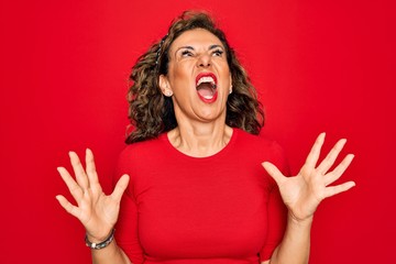 Middle age senior brunette woman wearing casual t-shirt standing over red background crazy and mad...