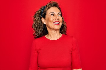 Middle age senior brunette woman wearing casual t-shirt standing over red background looking away to side with smile on face, natural expression. Laughing confident.