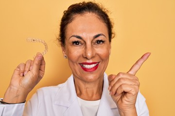 Middle age senior dentist woman holding clear aligner for teeth correction very happy pointing with...