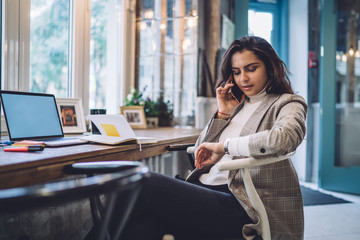 Caucasian female freelancer checking time on wearable wrist watch during time for remote work on blank laptop with copy space area for internet advertising, young woman talking via cellphone app