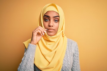 Young beautiful african american girl wearing muslim hijab over isolated yellow background mouth and lips shut as zip with fingers. Secret and silent, taboo talking