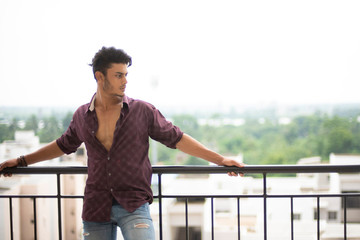 Portrait of an young and handsome brunette Bengali muscular man in casual shirt and jeans standing on a balcony in white urban background. Indian lifestyle.