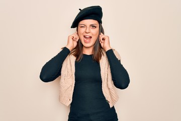 Young beautiful brunette woman wearing casual french beret over white background covering ears with fingers with annoyed expression for the noise of loud music. Deaf concept.