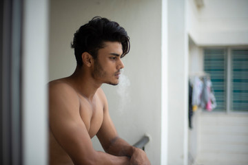 Fototapeta na wymiar Portrait of an young and handsome brunette Bengali muscular man in bare body smoking on a balcony in white urban background. Indian lifestyle.