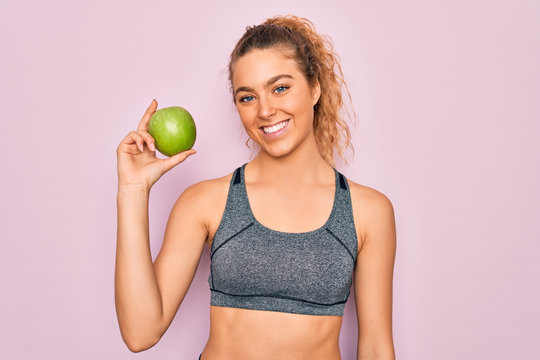 Young beautiful blonde sporty woman with blue eyes holding healthy green apple fruit with a happy face standing and smiling with a confident smile showing teeth