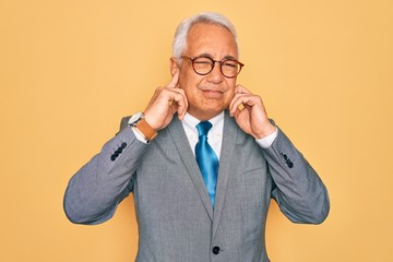 Middle age senior grey-haired handsome business man wearing glasses over yellow background covering ears with fingers with annoyed expression for the noise of loud music. Deaf concept.