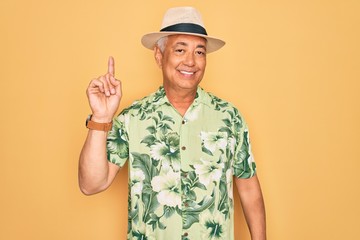 Middle age senior grey-haired man wearing summer hat and floral shirt on beach vacation showing and pointing up with finger number one while smiling confident and happy.