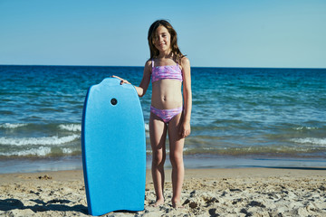 portrait of a girl facing the beach by the sea and holding a blue surfboard - obrazy, fototapety, plakaty