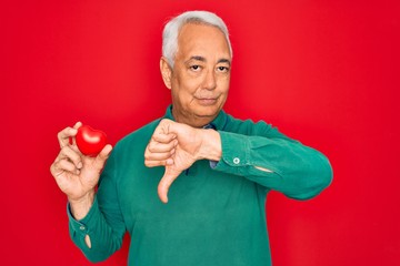 Middle age senior grey-haired man holding romantic red heart object over isolated red background...