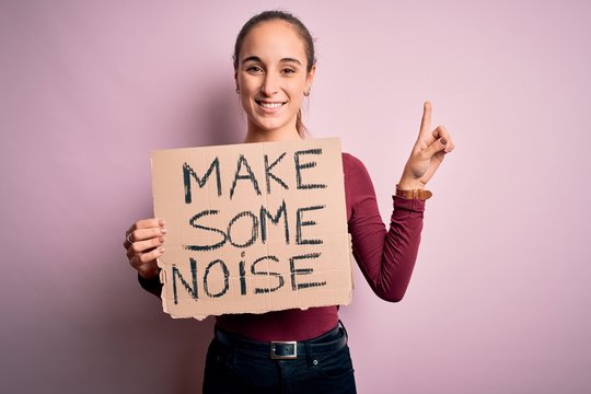 Beautiful activist woman holding banner with make some noise message over pink background surprised with an idea or question pointing finger with happy face, number one