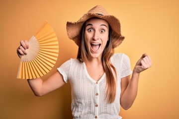 Young tourist woman on vacation wearing summer hat holding hand fan over yellow background screaming proud and celebrating victory and success very excited, cheering emotion