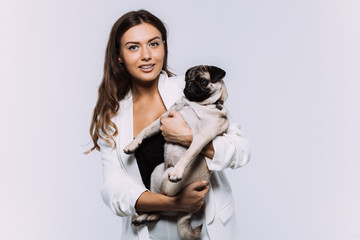 A laughing and smiling auburn haired woman in a white dress, is staring most lovingly at her cute pug, who calmly sits on the hands, gaining her undivided attention. Isolated white background.
