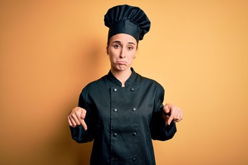 Young beautiful chef woman wearing cooker uniform and hat standing over yellow background Pointing down looking sad and upset, indicating direction with fingers, unhappy and depressed.