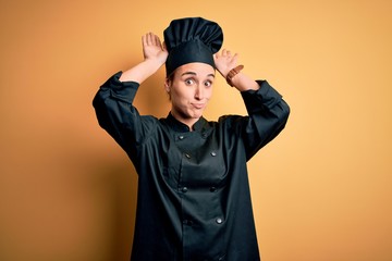 Young beautiful chef woman wearing cooker uniform and hat standing over yellow background Doing bunny ears gesture with hands palms looking cynical and skeptical. Easter rabbit concept.