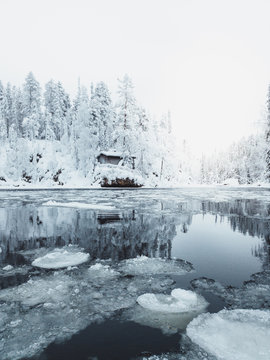 Kleine verschneite Holzhütte an einem Fluss mit Eisschollen im Vordergrund, umrundet von verschneiten Bäumen am Polarkreis