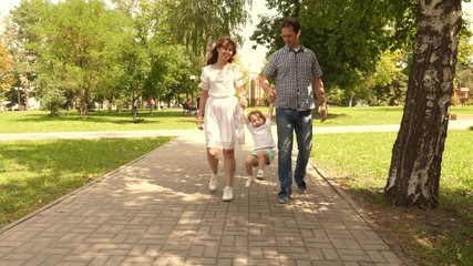 happy child plays with dad and mom holds their hands and jumps. little daughter jumping holding hands of dad and mom in the park. Family concept. Walking with small kid in nature.