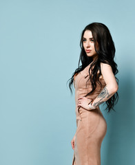 Brunette model in beige dress. There is watch on her tattooed hand. Standing sideways against blue studio background. Close up