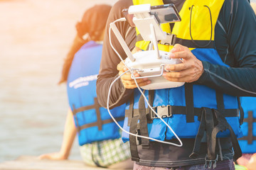 Fototapeta na wymiar Young man in blue and yellow life vest controlling a drone to take pictures of ocean coast with tourists while traveling on a raft in the sea. Hands holding drone remote controller.