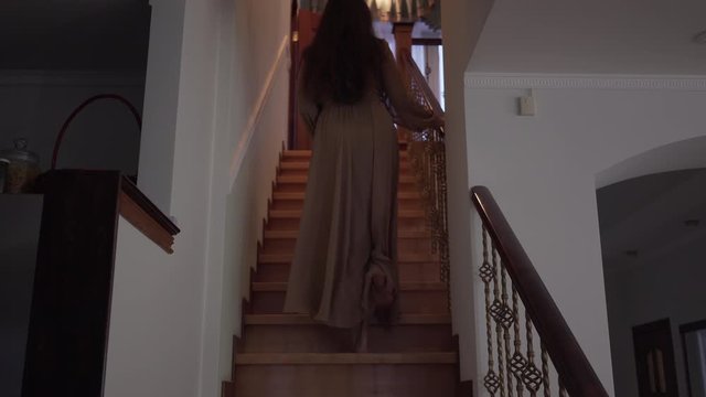 Back view of eerie woman walking up the stairs slowly and entering room in house. Brunette barefoot witch indoors. Magic, horror, nightmare, paranormal.