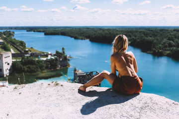 At a picturesque vantage point of Rummu quarry, a blond woman is standing in her bare feet, wearing a thigh high, auburn coloured dress, With her back turned away from the camera. Clear Water.