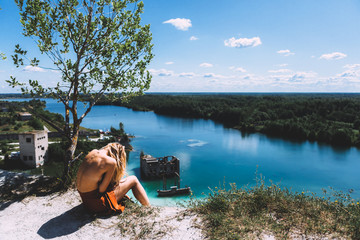 At a picturesque vantage point of Rummu quarry, a blond woman is sitting, wearing a thigh high, auburn coloured dress, With her back turned away from the camera. Clear Water.