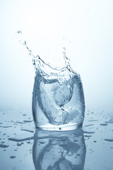A piece of ice falling in a glass of water and raising a big splash. Water wave and splatter on white