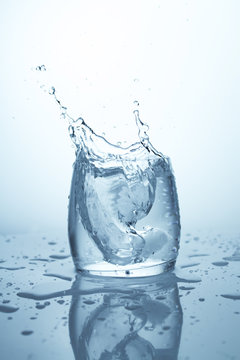 A Piece Of Ice Falling In A Glass Of Water And Raising A Big Splash. Water Wave And Splatter On White