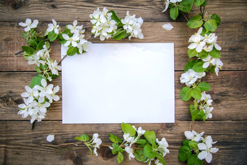 A branch of a blooming apple tree with white flowers and a sheet of paper on a wooden background, with a copy space