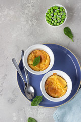 Two white ceramic ramekins with baked cheese souffle with green peas on a light grey table. Top view.