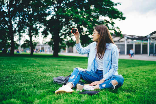 Young woman in casual wear using smartphone camera shooting video for blog sitting on green park on free time, female influencer taking selfie on mobile phone communicating with followers on vacations