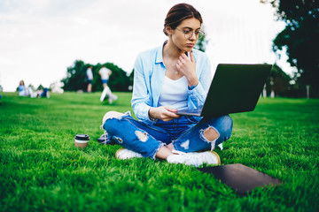 Serious pensive hipster girl in eyewear for vision protection reading latest news from web page while sitting on grass in park, young millennial woman using laptop computer for learning online