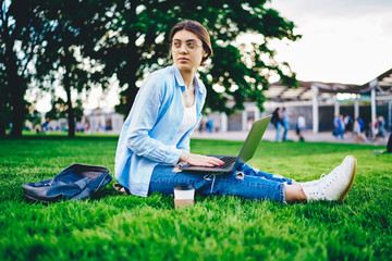 Serious caucasian female student sitting on college campus doing homework online via 4G connection, millennial young woman using laptop computer for publication in blog typing articles on rest in park