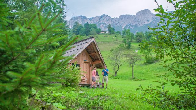 Adult people standing at a wooden cabin near a forest in the middle of green valley, with mountains in the distance.