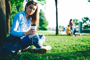 Portrait of caucasian woman recreating outdoors near tree listening playlist songs on smartphone and accessory, young teen millennial hipster girl using mobile phone app and sound earphones in park