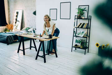 Portrait of happy cheerful female planning travel trip with paper map during free time in cozy apartment interior with blank pictures on wall for advertising text, woman in spectacles posing at home