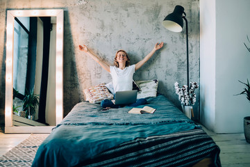 Enthusiastic female sitting on bed with hands up and using laptop in room