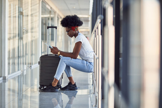 Young african american female passanger in casual clothes is in airport with baggage. Using phone